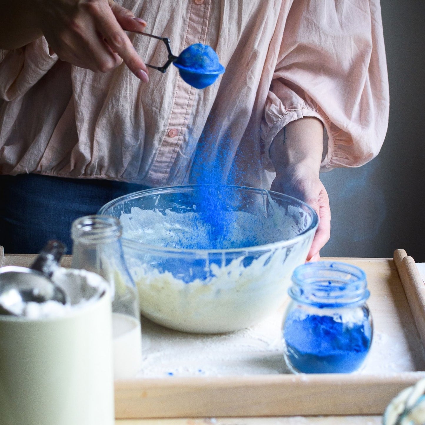 Person adding blue powder to a bowl of white substance on a wooden table.