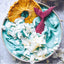 Blue smoothie bowl with a pineapple slice, mermaid tail, and coconut flakes on a gray surface.